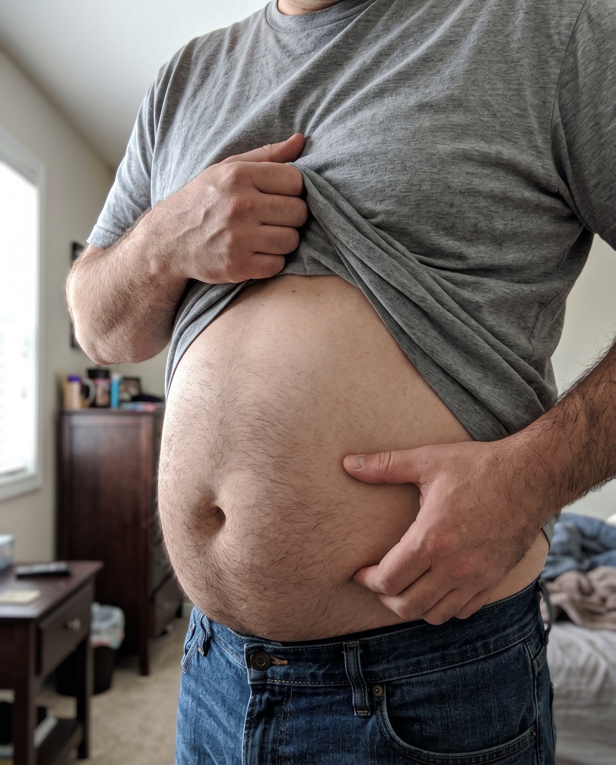 A man in his fifties lifting his shirt over a round belly in a bedroom